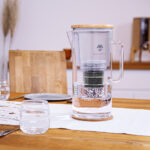 Eine Glas-Wasserfilterkanne mit Bambusdeckel und sichtbarer Filterkartusche steht gefüllt mit Wasser auf einem gedeckten Holztisch.