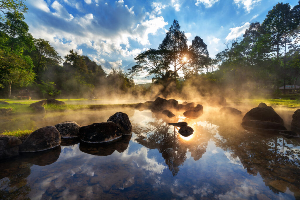 Eine natürliche, dampfende heiße Quelle bei Sonnenaufgang, deren klares Wasser den Himmel widerspiegelt und von üppiger Natur umgeben ist.