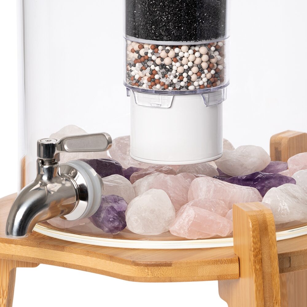 Detailansicht eines Glas-Wasserfilters mit mehrstufiger Filterkartusche und Edelsteinen wie Rosenquarz und Amethyst, stehend auf einem Bambussockel.