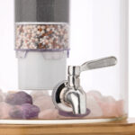 Nahaufnahme eines Edelstahl-Wasserhahns an einem Glas-Wasserspender, im Hintergrund eine mehrstufige Filterkartusche und am Boden Edelsteine.