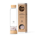 Edle Glas-Trinkflasche mit Bambusdeckel und separatem Fach, gefüllt mit einer Mischung aus Rosenquarz-, Bergkristall- und Amethyst-Edelsteinen, stehend neben der zugehörigen Kartonverpackung.