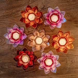 Sieben bunte Lotusblüten-Teelichthalter mit brennenden Kerzen, von oben auf einer Holzoberfläche arrangiert.