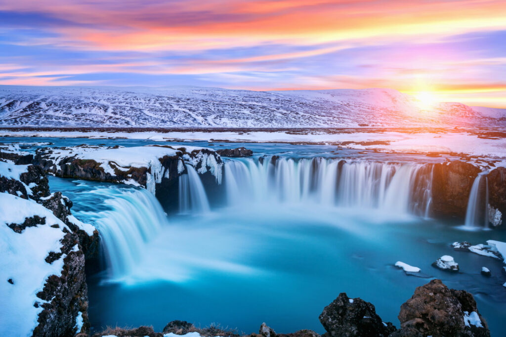 Der isländische Godafoss-Wasserfall, umgeben von einer unberührten Schneelandschaft bei Sonnenuntergang, symbolisiert die Kraft und Reinheit natürlichen Wassers.
