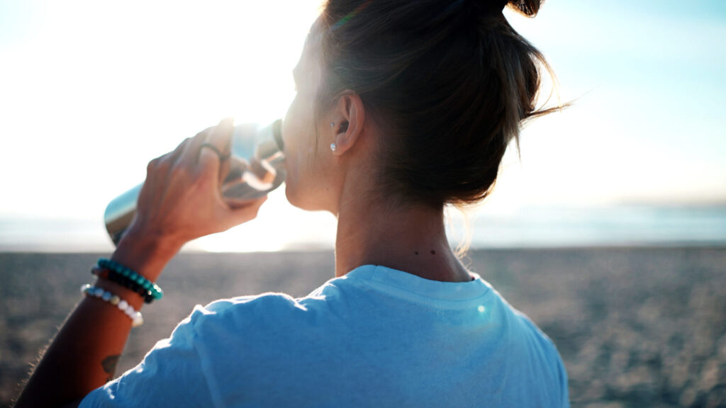 Rückansicht einer Frau, die bei Sonnenuntergang am Strand aus einer nachhaltigen Trinkflasche trinkt und ihren Körper nach dem Sport mit Flüssigkeit versorgt.
