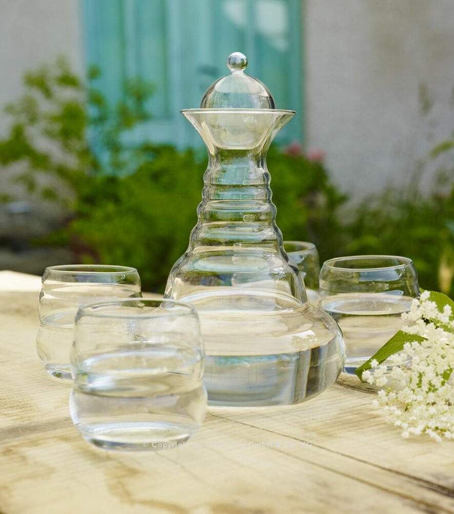 Eine formschöne Glaskaraffe und vier passende Trinkgläser, gefüllt mit reinem Wasser, stehen auf einem Holztisch in einem sommerlichen Garten.