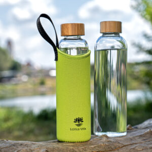 Zwei Glas-Trinkflaschen mit Bambusdeckel, eine davon in einer leuchtend grünen Neopren-Schutzhülle, stehen auf einem Baumstamm in der Natur.