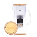 Glas-Wasserfilterkanne mit sichtbaren Filter-Schichten und einem Bambus-Deckel, daneben ein goldener Deckel mit eingraviertem Lebensblume-Muster.