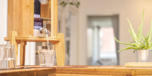 Ein Wasserfilter-Standgerät aus Bambus mit einem Zapfhahn aus Edelstahl befüllt ein Trinkglas mit frischem, klarem Wasser.