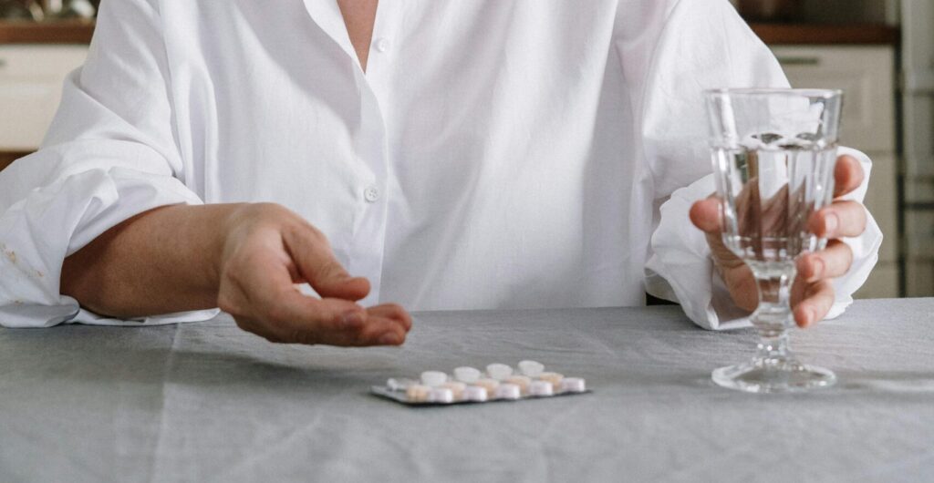 Eine ältere Frau in ihrer Küche hält ein Glas Wasser und eine Tablette in der Hand, um ihre Medikamente einzunehmen.