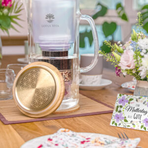 Eine Glas-Filterkanne mit einem goldenen Deckel, verziert mit der Blume des Lebens, steht auf einem liebevoll für den Muttertag gedeckten Tisch neben einer Grußkarte.
