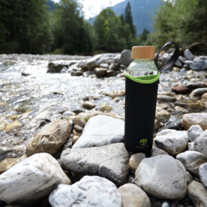 Eine Glas-Trinkflasche mit schwarzer Schutzhülle und Holzdeckel steht auf den Steinen eines klaren Gebirgsbachs im Allgäu.