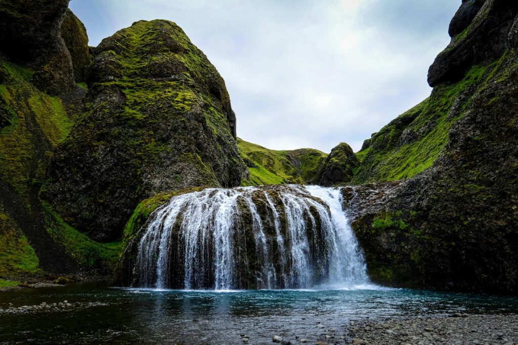 Ein breiter Wasserfall fließt in Kaskaden über moosbewachsene Felsen in einen ruhigen, klaren See.