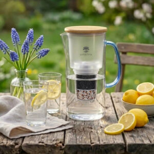 Ein Glas-Wasserfilterkrug mit Holzdeckel und blauem Griff steht auf einem Holztisch im Freien, umgeben von Gläsern mit Zitronenwasser und frischen Blumen.