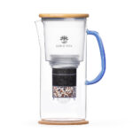 Ein Glas-Wasserfilterkrug mit blauem Henkel und Holzdeckel steht auf einem Korkuntersetzer und zeigt die Filterkartusche im Inneren.
